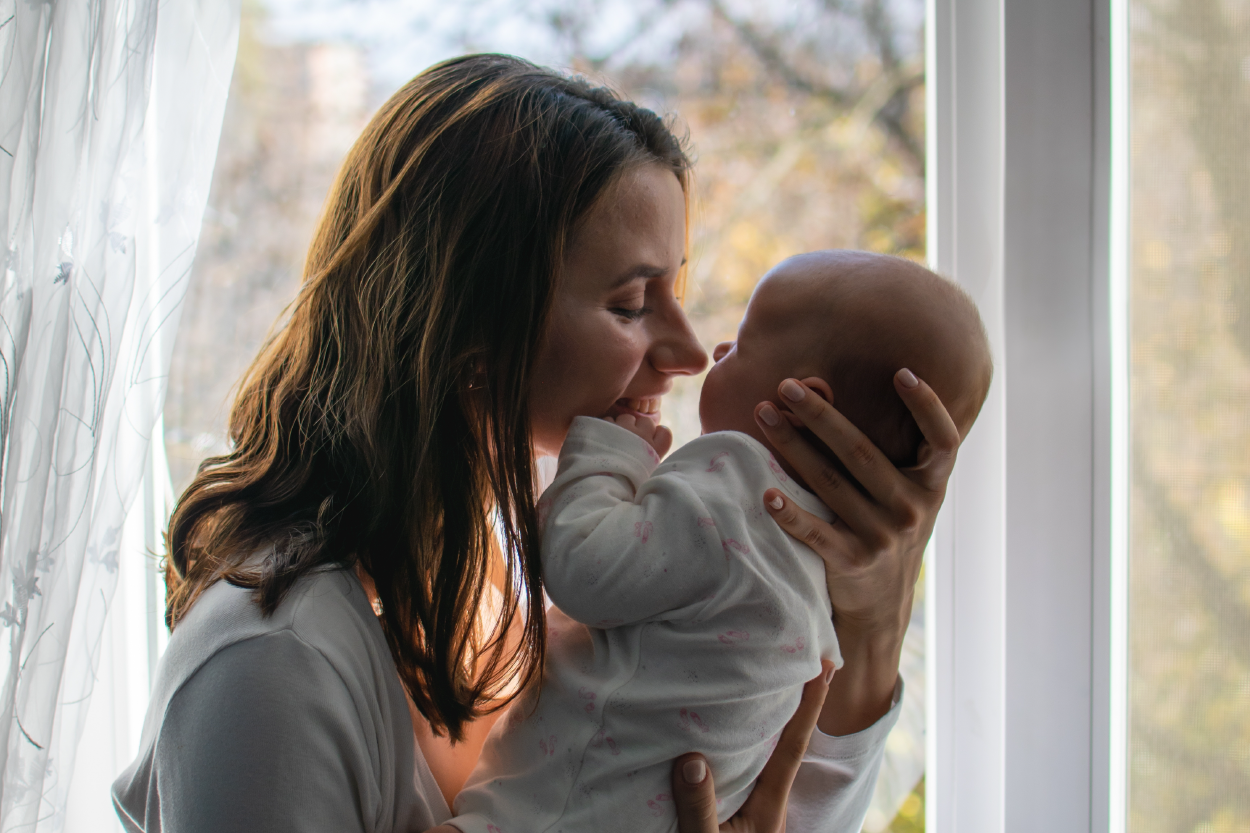 A woman standing in front of a window holds an infant up to her face. The infant's hand is on her chin and their noses almost touch.