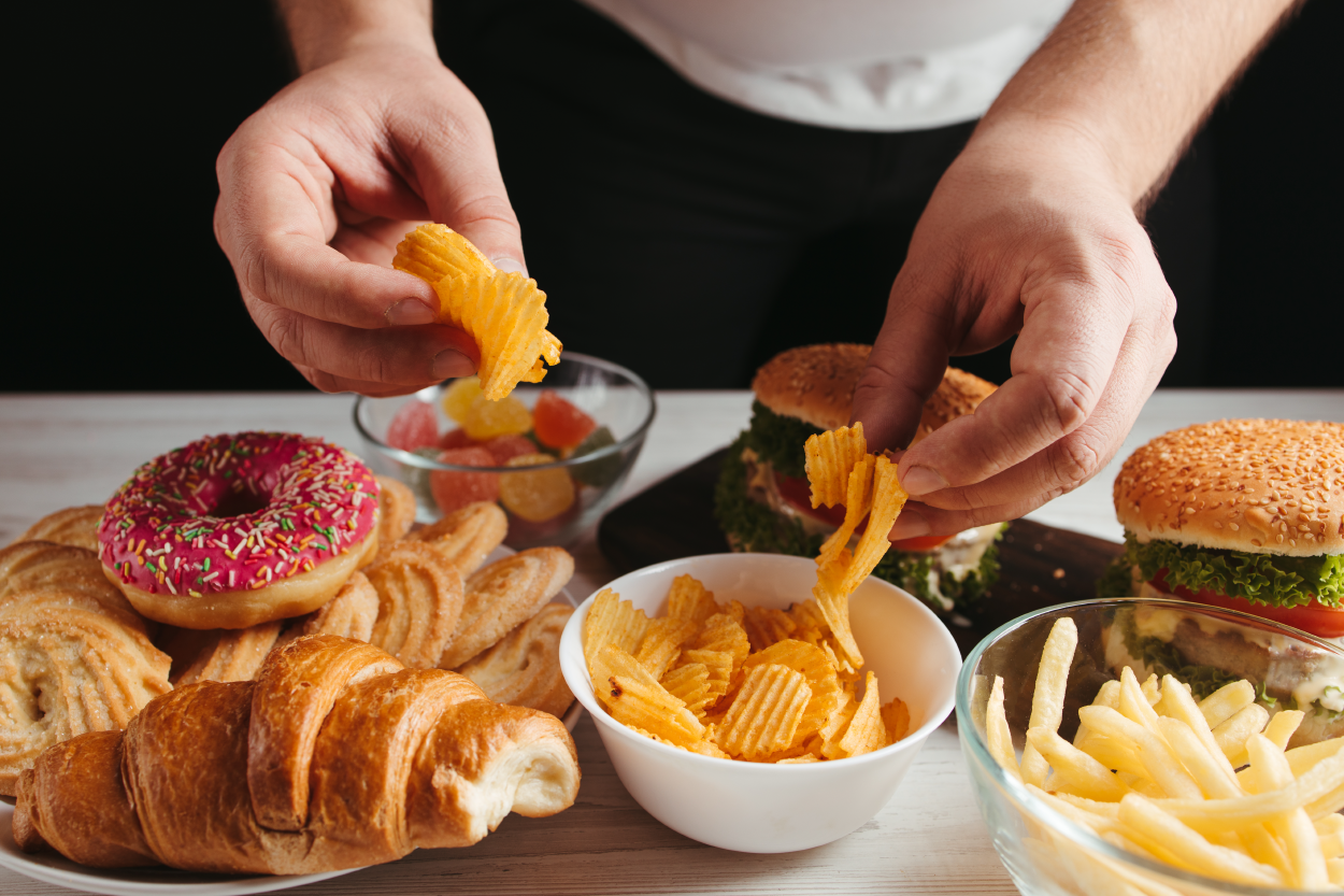 A close-up of someone eating; two hands are grabbing food from a table with pastries, hamburgers, chips, and fries