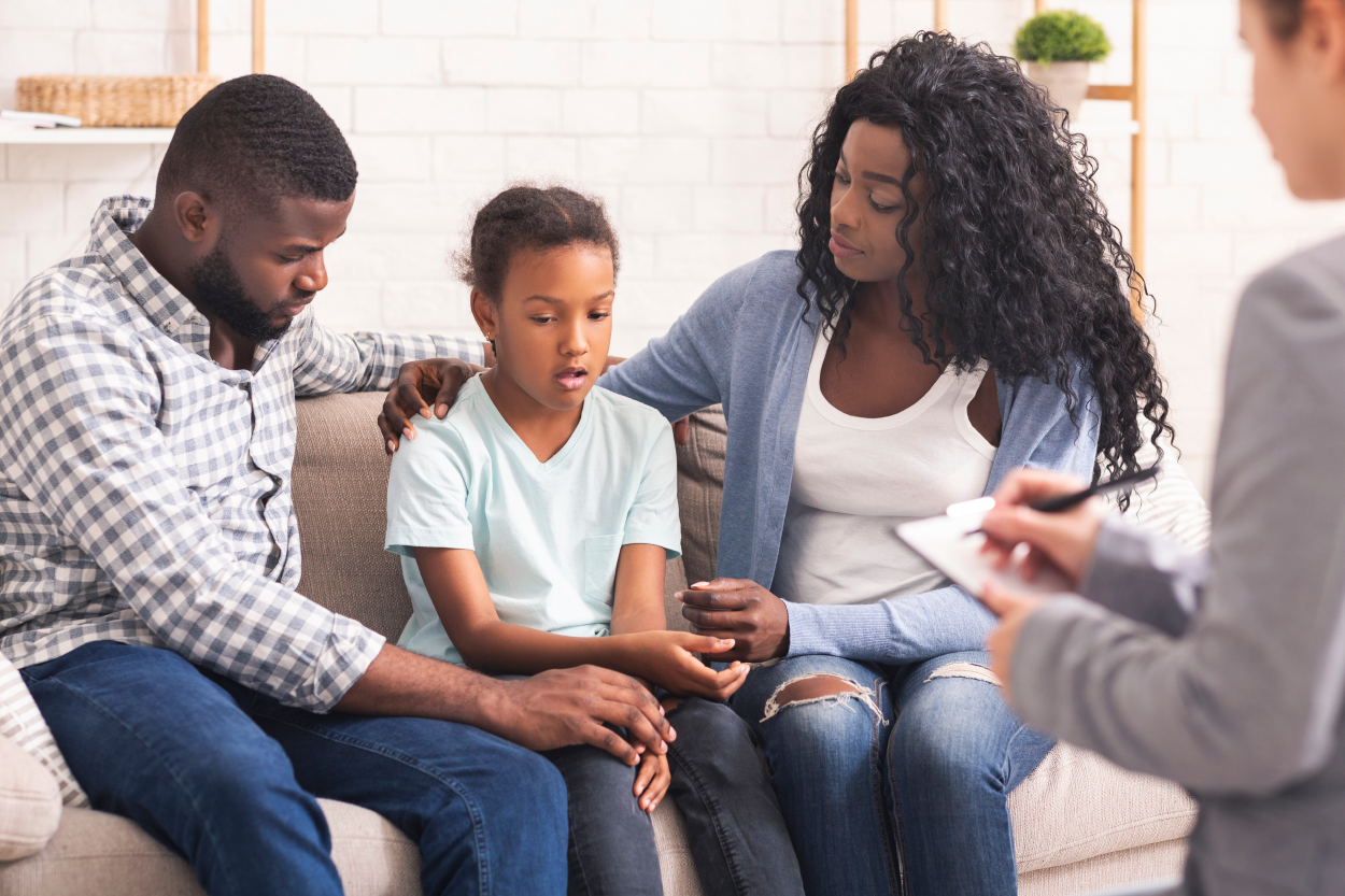 A young Black child sitting on a couch with her mother and father. The mother has her arm on the child's shoulder. A person sit across from them taking notes.