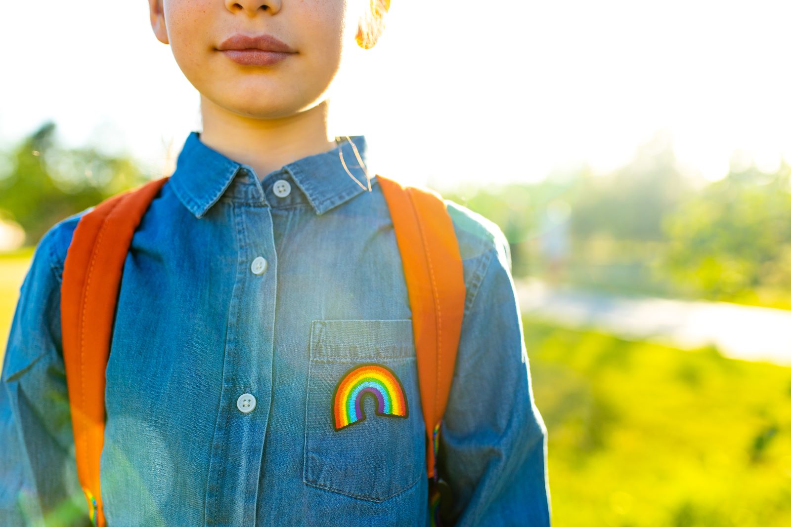 child in denim t-shirt with rainbow symbol, wearing an orange backpack outdoor, standing outside
