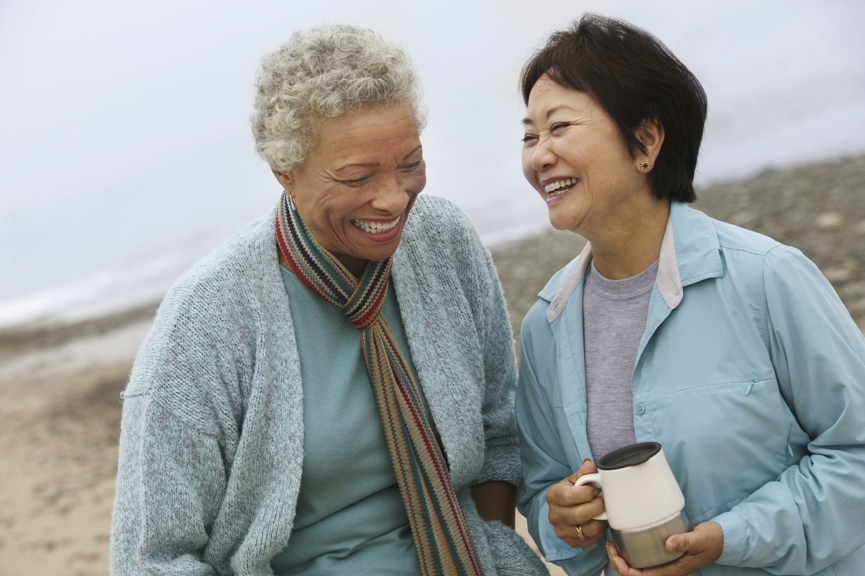 Two older women laughing; walking on the beach; one holding coffee; adult friendship