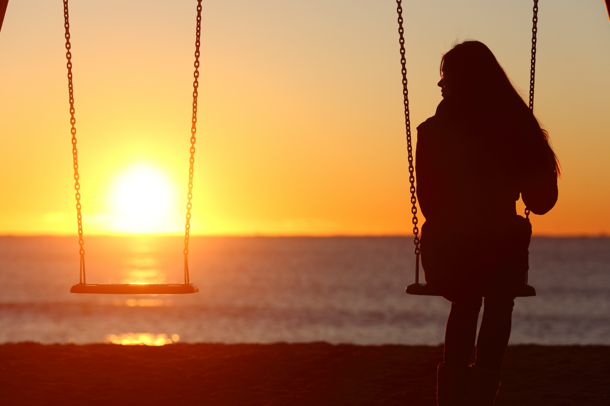 An adult sitting in a swing looking back at a sunset over the ocean