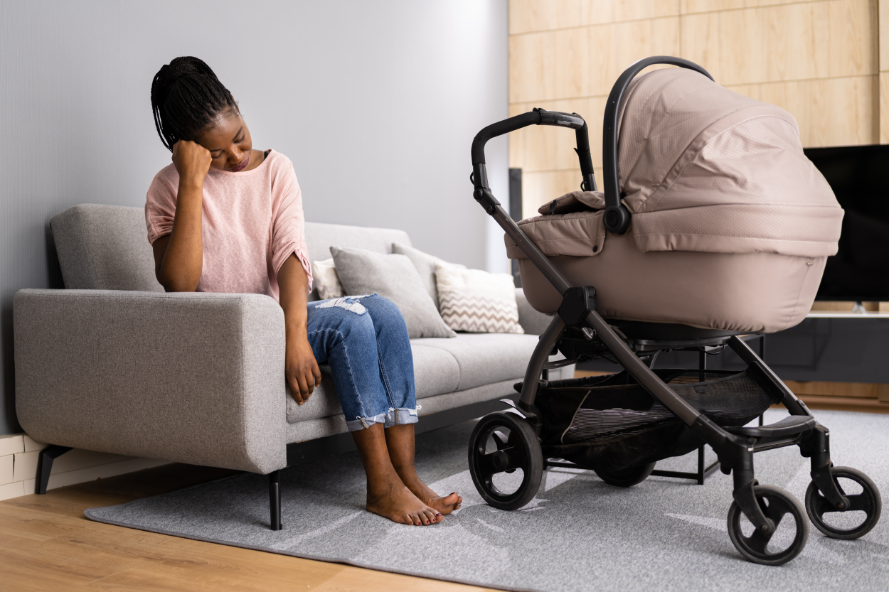A Black mother sits on a sofa with a baby stroller in front of her. She is looking down. She looks tired and stressed.