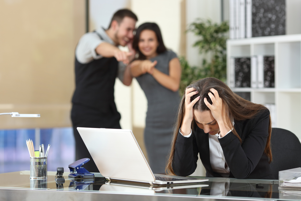 A woman being bullied at work. Two coworkers are pointing and laughing at her. She has her hands on her hand, looking down, appears stressed.