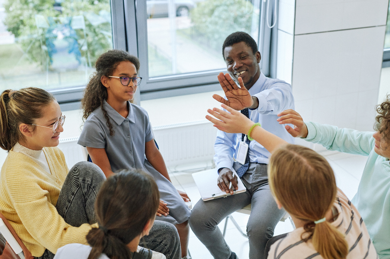 A counselor sits in a circle with a group of kids. The counselor has a notepad and pen and is giving two of the kids high fives.