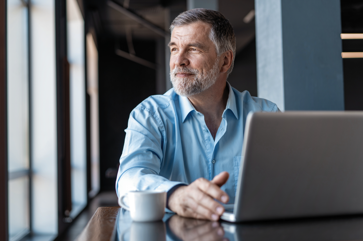 older white man sitting at a table with a laptop and coffee cup, looking out the window