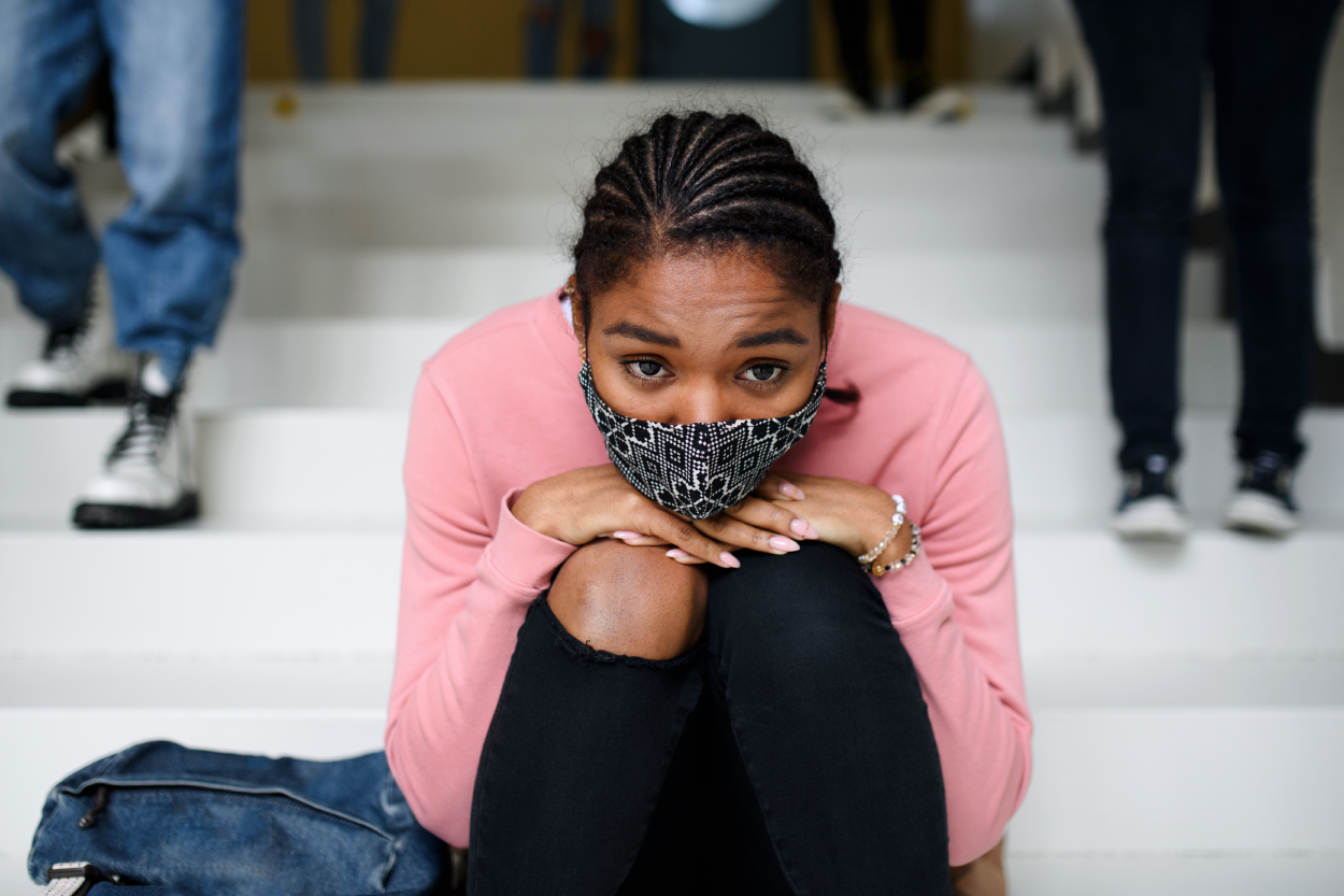 A teenager wearing a mask sits on stairs with her chin and hands resting on her knees. The teenager is looking straight head into the camera. A bookbag sits beside the teenager.